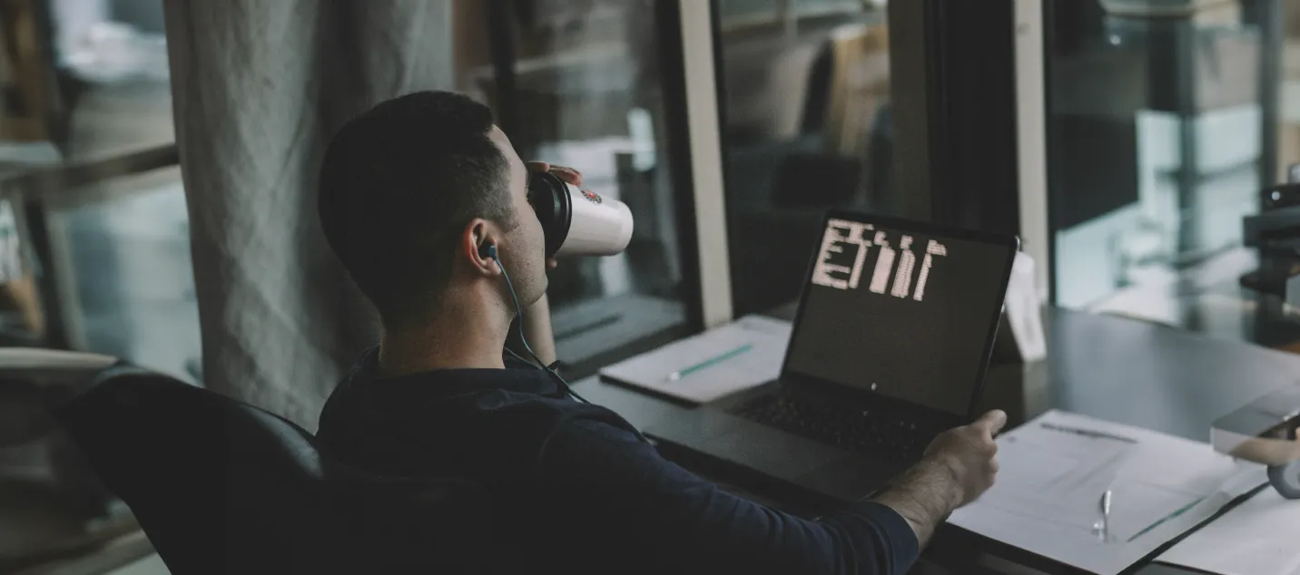 Image of a man looking at his laptop on his desk and sipping coffee.